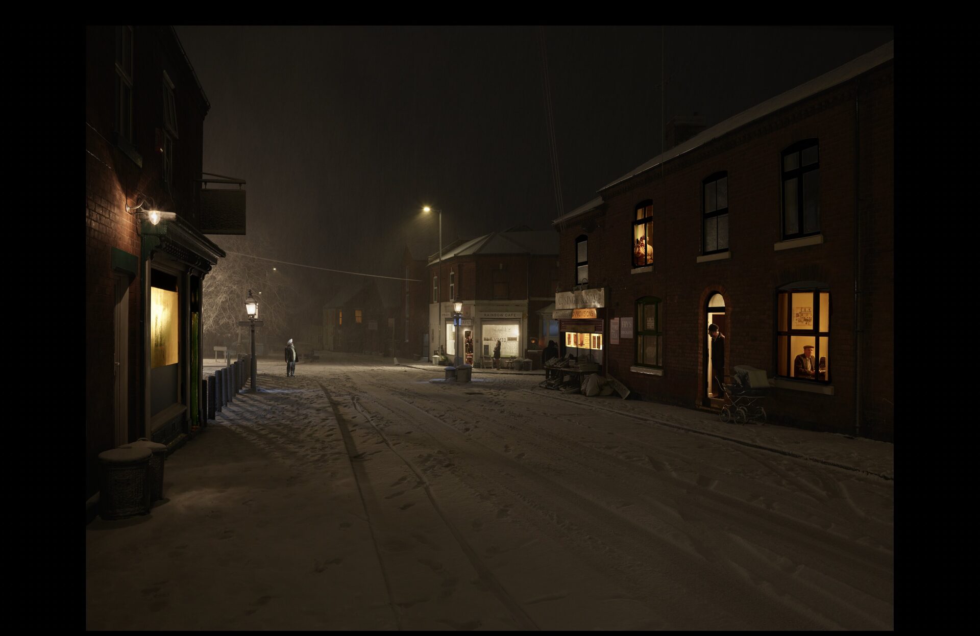 A snow-covered Black Country street at night. Terraced houses with warm lit windows line both sides. Streetlamps glow through the falling snow. A solitary figure walks away down the centre of the empty road. Shopfronts and a church tower are visible in the haze.