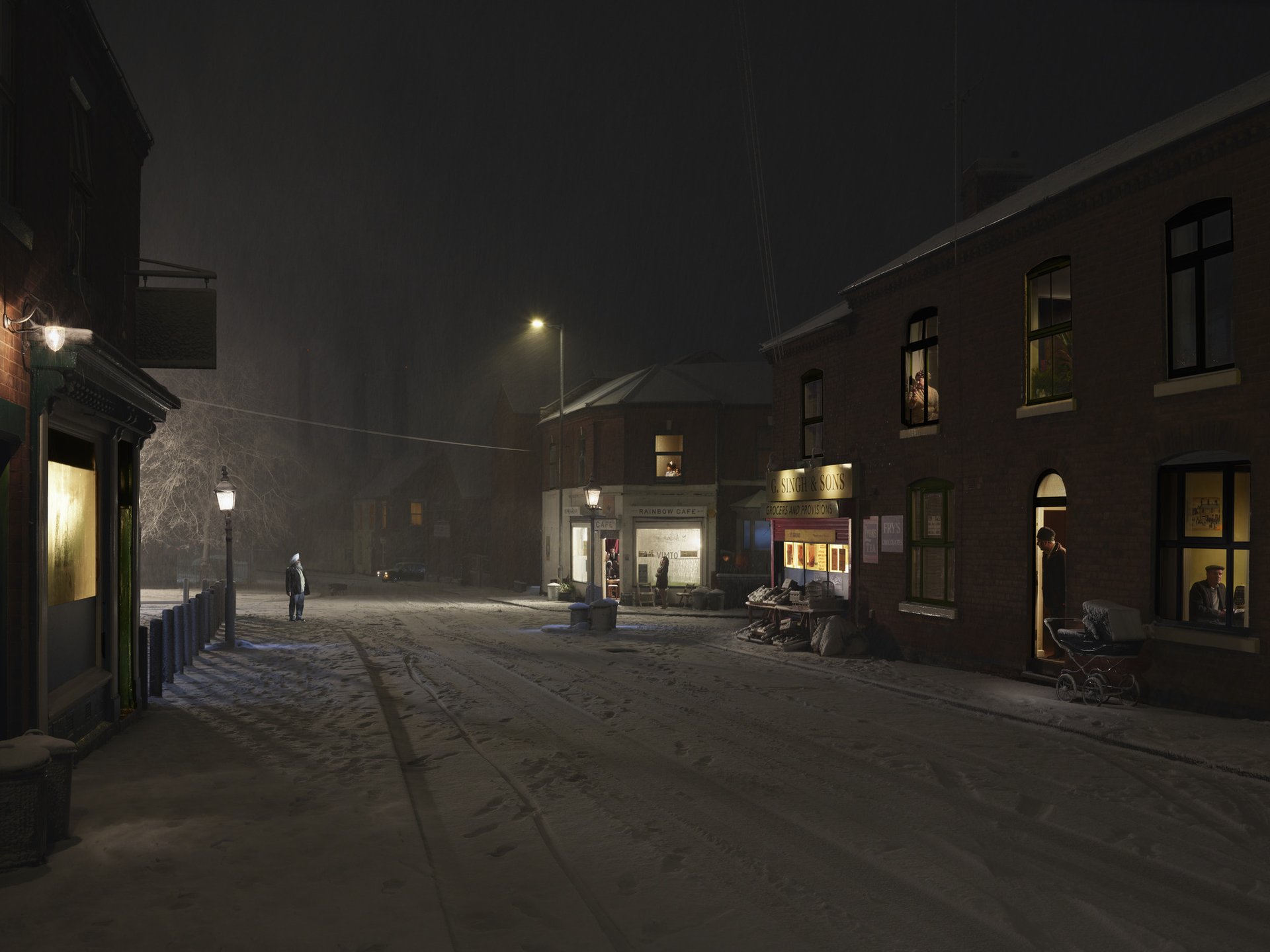 A snow-covered Black Country street at night. Terraced houses with warm lit windows line both sides. Streetlamps glow through the falling snow. A solitary figure walks away down the centre of the empty road. Shopfronts and a church tower are visible in the haze.