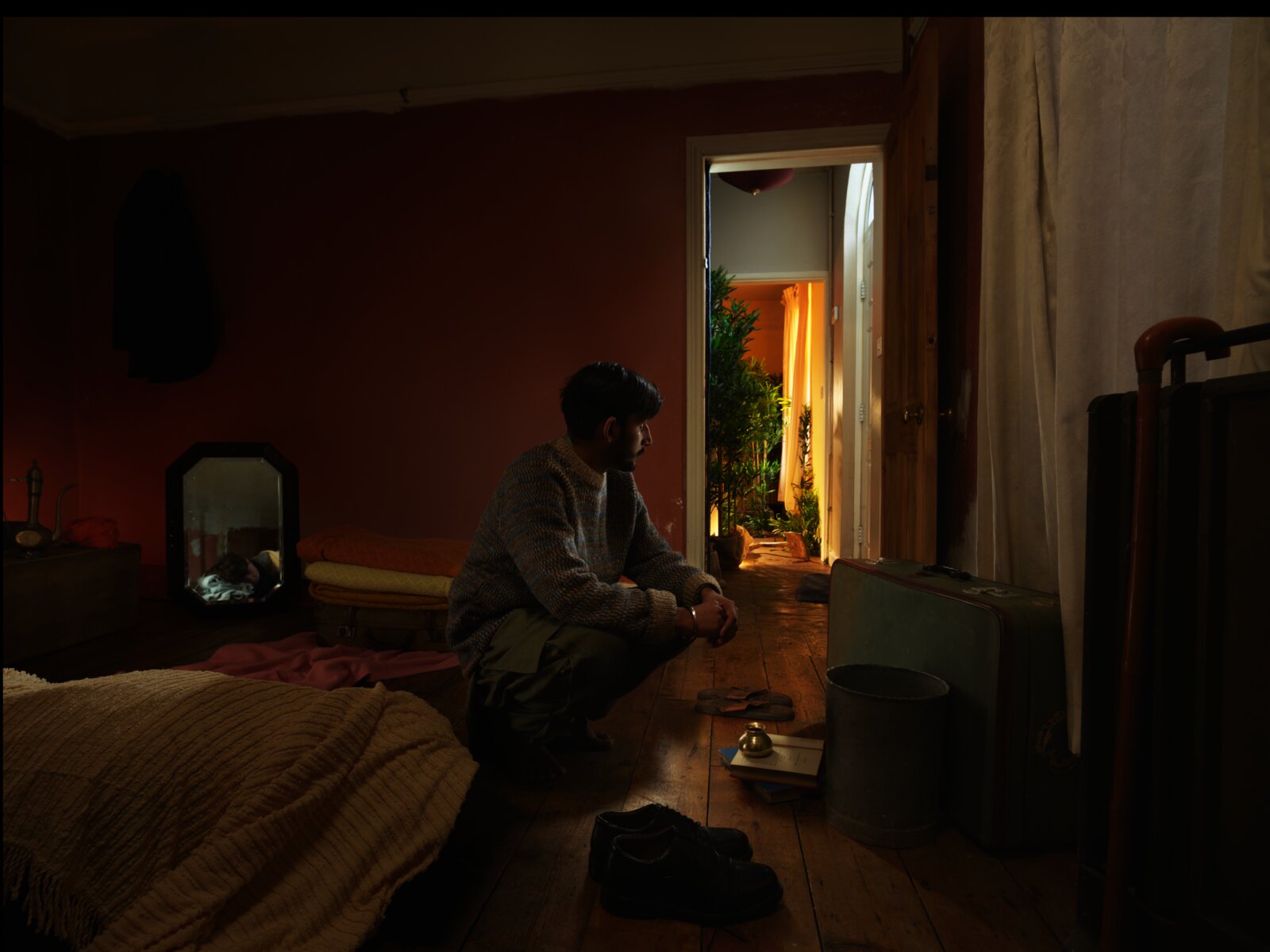 A young man sits on bare floorboards in a dimly lit room, looking towards a doorway filled with golden light and dense foliage. A suitcase and pair of shoes rest on the floor beside him. A bed and mirror are visible in the shadows.