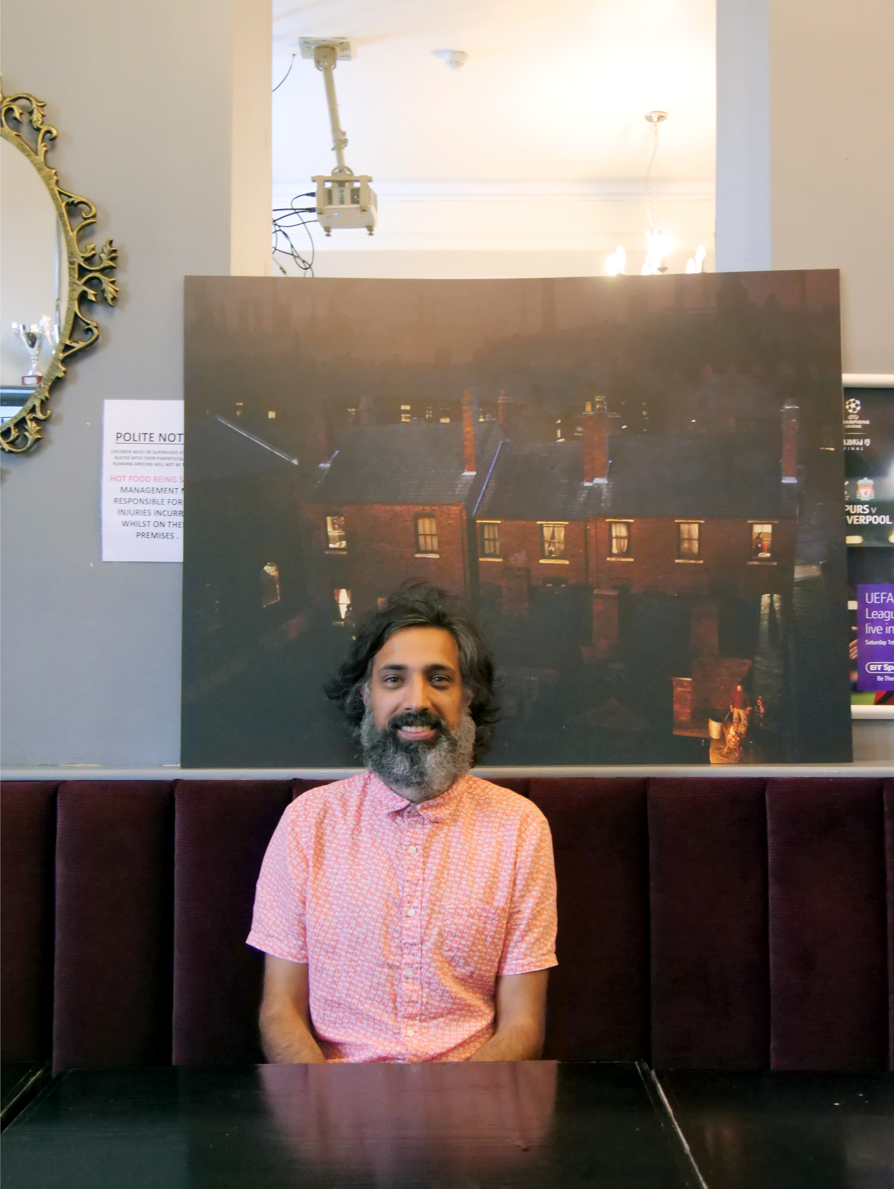 Billy Dosanjh, Artistic Director of Reimagining Industrial Migratory Stories CIC, seated in front of one of The Exiles photographic prints.