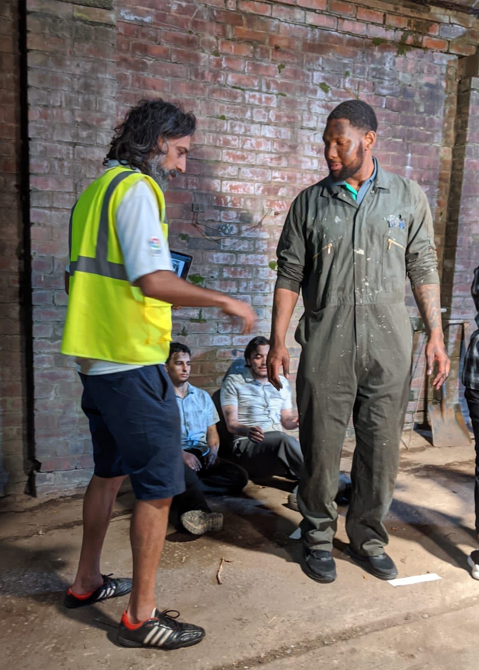 Director positioning a performer in factory overalls against a brick wall on the Furnace Men set