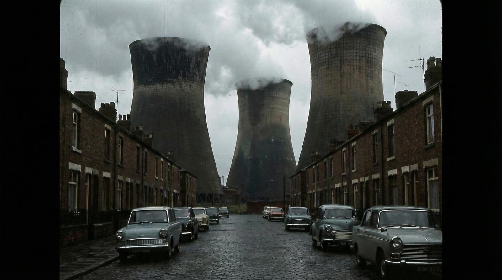 Industrial cooling towers dominating a row of Victorian terraced houses in wet evening light — the setting of The Exiles